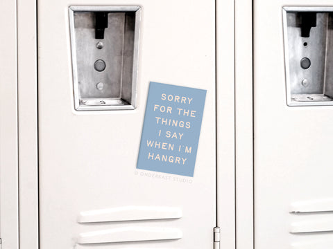 A blue Hangry Refrigerator Magnet by Onderkast Studio, featuring vibrant colors and the message “Sorry for the things I say when I’m hangry,” is displayed on a white locker with adjacent vented lockers and locks visible.