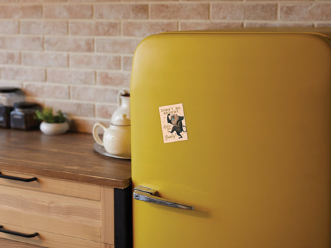 A retro yellow fridge with an Onderkast Studio Shake Ya Booty Baboon Magnet on its door stands in a cozy kitchen featuring wooden cabinets, jars, and a white teapot on the counter against a brick backsplash.