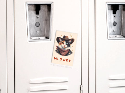 A white locker door with the Onderkast Studio Meowdy Cowboy Cat Refrigerator Magnet attached, showing a postcard of a cat in a cowboy hat and bandana with MEOWDY beneath the illustration.