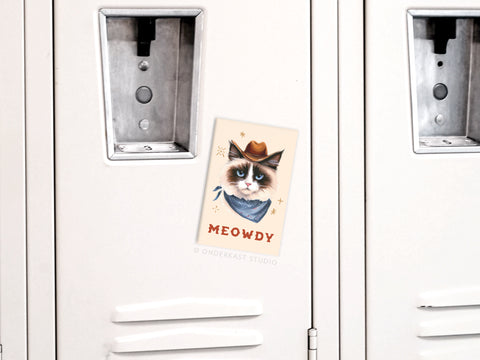 A locker decorated with a postcard shows a grumpy cat in a cowboy hat and bandana, captioned “MEOWDY”—the perfect inspiration for the Meowdy Cowboy Cat Refrigerator Magnet by Onderkast Studio.