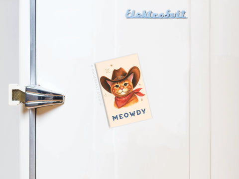 A white fridge with a metal handle showcases the Meowdy Cowboy Cat Refrigerator Magnet by Onderkast Studio—depicting a cat in a cowboy hat and bandana—and a light blue Elektrobit magnet, adding charm to this whimsical display.
