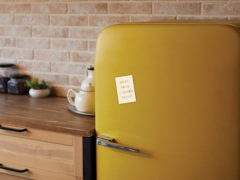 A yellow retro fridge in a kitchen with a wooden counter, jars, and teapot features the Onderkast Studio Make Good Choices Today Refrigerator Magnet against a brick-patterned wall.