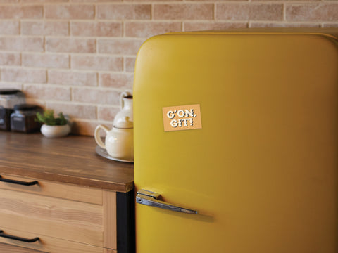A retro yellow fridge featuring the Onderkast Studio G’on Git Refrigerator Magnet brings Southern charm to a cozy kitchen with brick walls, wooden counters, and jars in the background.