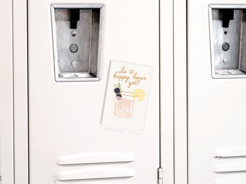 The Is It Happy Hour Yet Cocktail magnet by Onderkast Studio, featuring a chic cocktail illustration with a lemon slice and catchy text, is displayed on a white metal refrigerator alongside vibrantly colored surfaces.