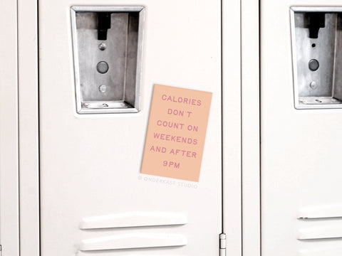 A beige locker displays the Onderkast Studio Calories Don’t Count Refrigerator Magnet, which reads, CALORIES DON’T COUNT ON WEEKENDS AND AFTER 9PM. Nearby lockers stay closed, each featuring sleek silver handles.