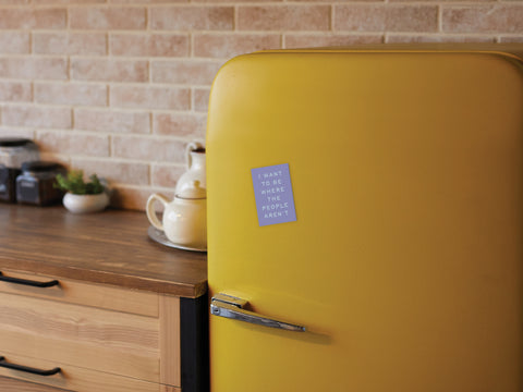 A yellow retro-style fridge in a cozy kitchen sports the Onderkast Studio “Be Where the People Aren’t” magnet, surrounded by wooden cabinets and a brick backsplash.