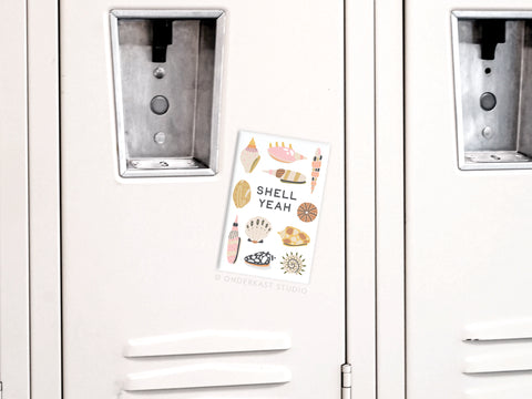 A white locker door displays the Onderkast Studio Shell Yeah Refrigerator Magnet, which holds a vibrant postcard featuring illustrated seashells and bold beach-inspired text.