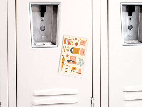 A white metal locker with a colorful autumn-themed poster showcasing socks, a book, tea, leaves, and an umbrella. Add extra charm by pairing it with the Cozy Vibes Refrigerator Magnet from Onderkast Studio.