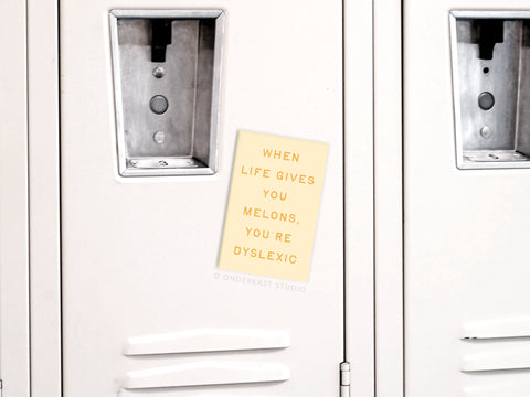 The Life Gives You Melons Refrigerator Magnet by Onderkast Studio, in beige with bold uppercase text, pops against a gray locker, humorously stating WHEN LIFE GIVES YOU MELONS, YOURE DYSLEXIC.