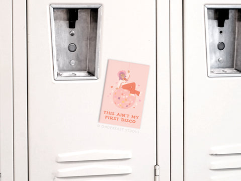 The Ain’t My First Disco Refrigerator Magnet by Onderkast Studio, featuring a woman on a disco ball and bold text, adds a pop of vibrant style to a light gray locker.