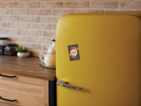 A cozy kitchen with a wooden countertop and brick wall features a retro yellow fridge adorned with the Onderkast Studio Yikes Fortune Teller Refrigerator Magnet, alongside a white teapot and jars for a whimsical touch.