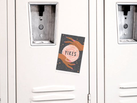 A Yikes Fortune Teller Refrigerator Magnet by Onderkast Studio features two illustrated hands holding a pink crystal ball with “YIKES” inside, set against a starry dark background—a whimsical design for fans of fortune teller style.