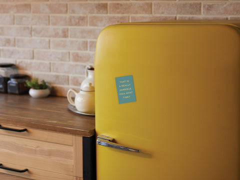 A retro yellow refrigerator features the Onderkast Studio Really Horrible Idea Refrigerator Magnet in blue on its door, set in a cozy kitchen with a wooden countertop, teapot, jars, and a brick backsplash.