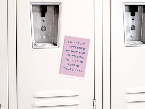 A pink note on a white locker reads, I’m pretty impressed by how bad I’m willing to look in public these days. The Willing to Look Bad In Public Refrigerator Magnet by Onderkast Studio adds a playful touch to your space. Nearby lockers are visible.