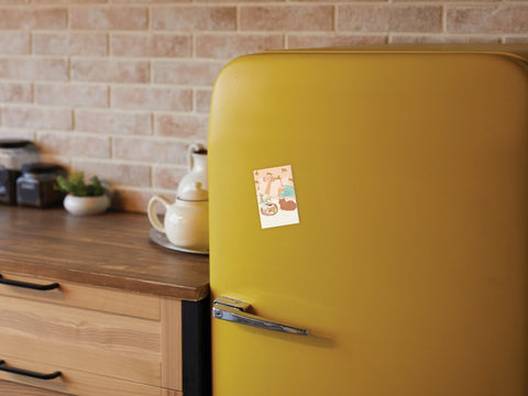 A yellow retro-style refrigerator features the Onderkast Studio Rise & Wine Refrigerator Magnet holding a note, standing in a kitchen with a wooden countertop, teapot, jars, and a brick backsplash.