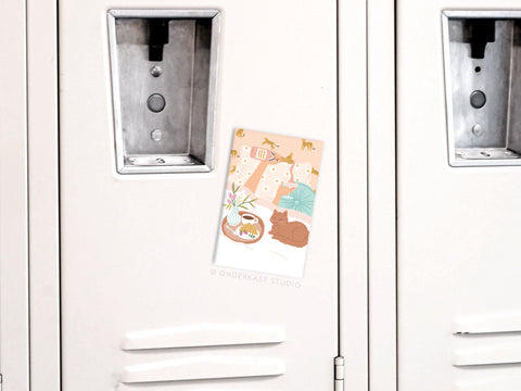 A white locker door displays a colorful cat-themed postcard, secured with the Rise & Wine Refrigerator Magnet by Onderkast Studio—featuring playful cat and plant illustrations, perfect for adding cheer to your kitchen.