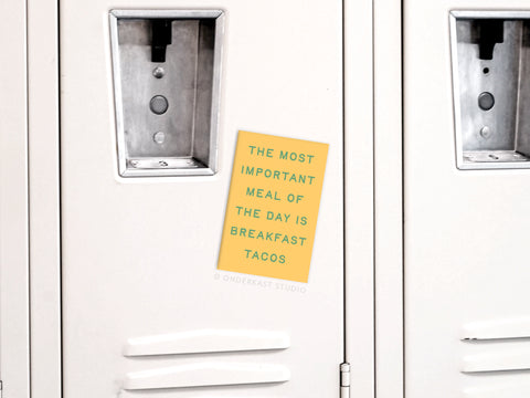 A yellow Breakfast Tacos Refrigerator Magnet by Onderkast Studio on a white locker reads, The most important meal of the day is breakfast tacos. The locker, ideal for kitchen decor, features a metal lock above the magnet.