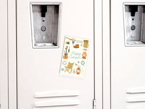 A white locker displays the Onderkast Studio Happy Camper Refrigerator Magnet, decorated with vibrant camping icons—a guitar, tent, backpack, lantern, campfire, and mug—ideal for outdoor lovers’ fridges or lockers.