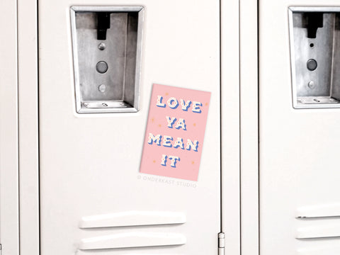 A Love Ya Mean It Refrigerator Magnet by Onderkast Studio, featuring vibrant pink colors and star accents, decorates a white locker among a row of lockers. The notes decorative lettering stands out beautifully.