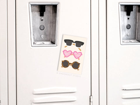 A white locker with a postcard showing three colorful sunglasses illustrations and an Onderkast Studio Sunglasses Refrigerator Magnet for a stylish magnetic display.