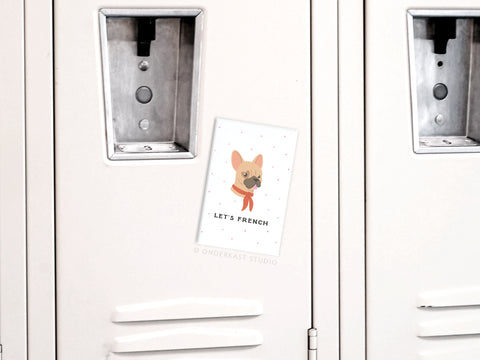 A gray locker features a postcard with a cartoon French bulldog and LETS FRENCH! attached by the Let’s French Frenchie Refrigerator Magnet from Onderkast Studio—an adorable gift for any dog lover.