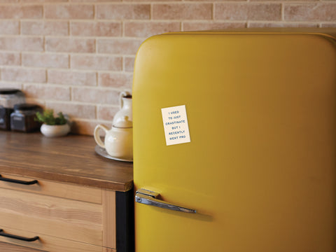 A yellow retro-style fridge features the Crastinater Pro Refrigerator Magnet by Onderkast Studio, displayed in a cozy kitchen with wooden countertops, a teapot, and jars against a beige brick backsplash.