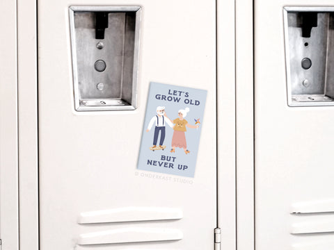 A Let’s Grow Old But Never Up Refrigerator Magnet by Onderkast Studio, featuring a cartoon elderly couple, is attached to a white locker.