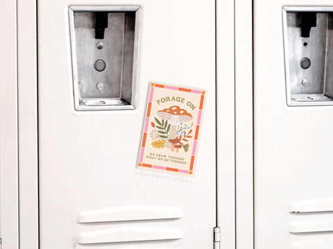 A vibrant Forage On Mushrooms Refrigerator Magnet by Onderkast Studio, featuring earthy illustrations and the phrase “FORAGE ON – WE GROW THROUGH WHAT WE GO THROUGH,” is attached to a white locker.