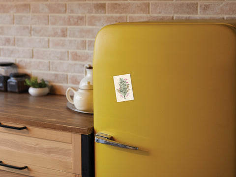 A yellow retro-style fridge with a small botanical postcard and the Good Vibes Smudge Stick Refrigerator Magnet by Onderkast Studio on its door sits in a kitchen with a wooden counter, jars, teapot, and brick backsplash.