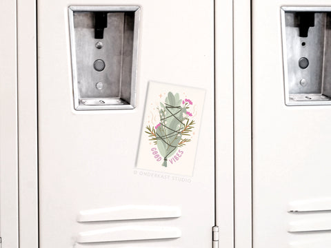 A white locker displays the Onderkast Studio Good Vibes Smudge Stick Refrigerator Magnet—a colorful cactus and flowers sticker—centered between two locks, adding bright energy to the space.