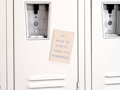 A beige “If I Stir It Then It’s Homemade” refrigerator magnet by Onderkast Studio, with blue text, is displayed on a white locker door between two metal handles, channeling kitchen confidence.