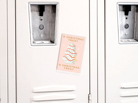 The O Christmas Treat Refrigerator Magnet by Onderkast Studio features vibrant pastel colors and a Christmas tree illustration with “Christmas Treat,” attached to a white metal locker.