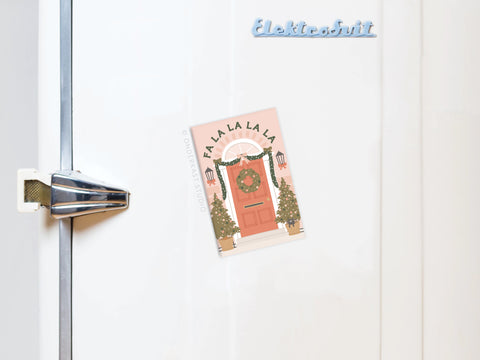 A white fridge with a chrome handle is festively decorated using Onderkast Studio’s Fa La La Christmas Door Refrigerator Magnet, which features an illustrated door with a wreath and “FA LA LA LA LA” design, perfect for holiday cheer.