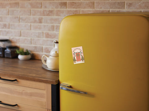 A yellow retro-style fridge features the Fa La La Christmas Door Refrigerator Magnet by Onderkast Studio holding a postcard; nearby, a teapot and jars rest on a wooden countertop with a brick backsplash in the background.