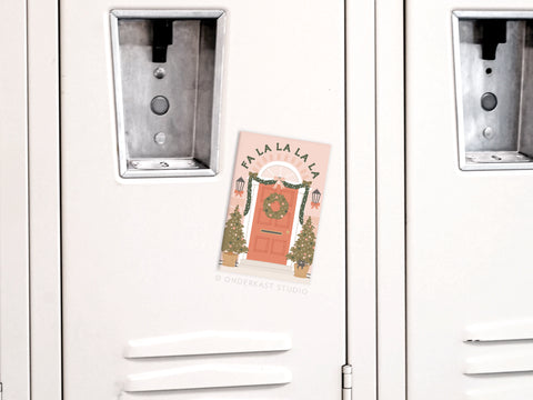 The Fa La La Christmas Door Refrigerator Magnet by Onderkast Studio attaches a holiday card with a red wreath-adorned door and festive trees to a white locker. The card reads FALA LA LA LA, creating a charming festive display.