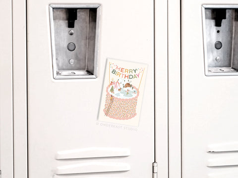 The Merry Birthday Cake Refrigerator Magnet by Onderkast Studio, featuring a festive cake design and cheerful message, is displayed on a light metal locker near the lock slot and handle, with small holiday accents adding charm.