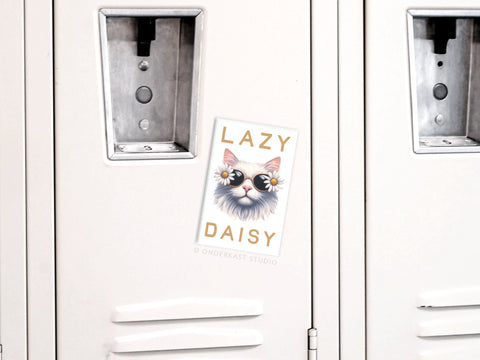 A white locker displays the Onderkast Studio Lazy Daisy Cat Refrigerator Magnet, featuring a cat in daisy sunglasses and Lazy Daisy text, adding playful feline charm.