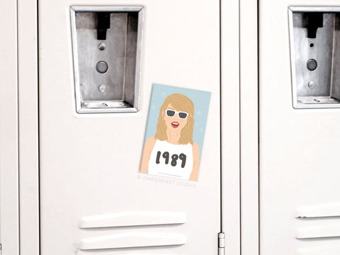 The Onderkast Studio TS 1989 Refrigerator Magnet features a cartoon woman with blonde hair, white sunglasses, and a 1989 shirt on a light blue, starry background—perfect for fans to decorate lockers or fridges.