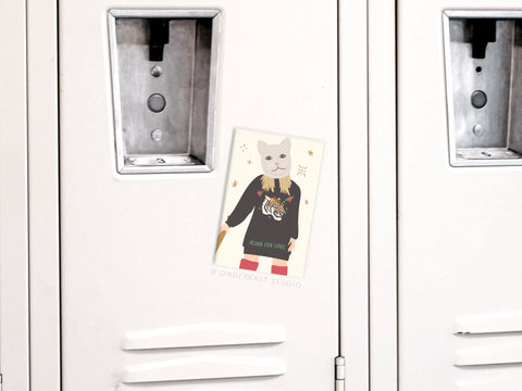 A white locker displays the TS Reputation Refrigerator Magnet by Onderkast Studio and a postcard with an illustrated cat in a dark sweater featuring a tiger design and BLIND FOR LOVE text on a star-sprinkled light background.