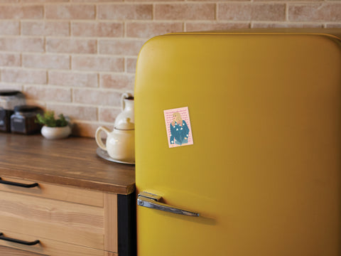 A cozy kitchen with wooden countertops, jars, a teapot, and a plant features a retro yellow refrigerator adorned with the Onderkast Studio TS Lover Refrigerator Magnet, set against a tan brick backsplash.