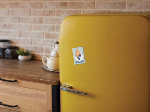 A retro yellow fridge is adorned with a TS Midnights Refrigerator Magnet by Onderkast Studio, showing an illustrated woman in winter clothes amid a cozy kitchen with brick walls, wooden counters, a teapot, jars, and celestial accents.