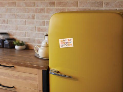 A retro yellow fridge with an Onderkast Studio Howdy Howdy Howdy Refrigerator Magnet, printed in the USA, stands in a kitchen with wooden cabinets, glass jars, a teapot, and a light brown brick backsplash.