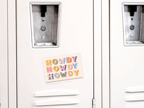 A white locker door displays the Onderkast Studio Howdy Howdy Howdy Refrigerator Magnet—made in the USA—which repeats “HOWDY” three times in orange, pink, and blue. The locker has a metallic handle and a mail slot above the magnet.