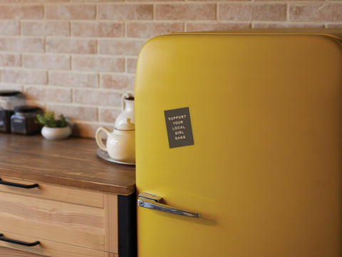 In a cozy kitchen with wooden cabinets and a brick backsplash, an Onderkast Studio Support Your Local Girl Gang magnet decorates a yellow retro fridge, surrounded by a teapot, cups, and jars that celebrate strong women.