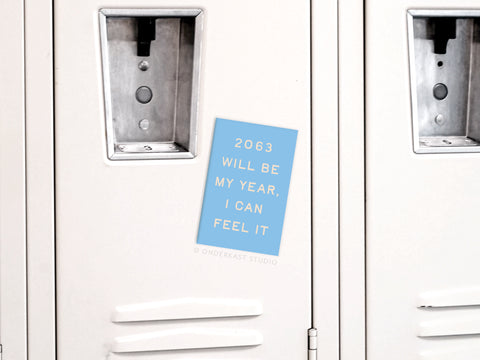 The Onderkast Studio 2063 Will Be My Year Refrigerator Magnet features a vibrant blue design and futuristic style, shown on a white locker with other lockers on each side.