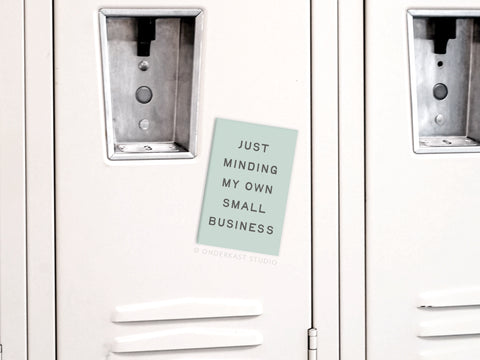 The Just Minding My Own Small Business Refrigerator Magnet by Onderkast Studio, featuring light blue design, is displayed on a white locker door between two other lockers—perfect for entrepreneurs on their business journey.