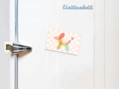 A white fridge with a metallic handle displays a postcard of a balloon dog, held by the Balloon Animal Refrigerator Magnet from Onderkast Studio, and features a blue Elektrobit magnet on the checkered background.