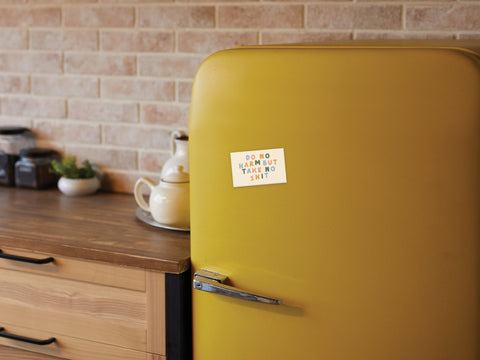 A yellow retro fridge in a cozy kitchen displays the Do No Harm But Take No Shit Refrigerator Magnet by Onderkast Studio, printed in the USA. The space features a wooden countertop and a brick backsplash.