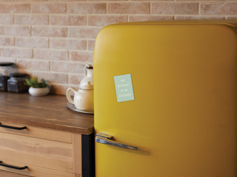 A retro yellow fridge features the Onderkast Studio No Bitchin’ In My Kitchen Refrigerator Magnet next to a wooden countertop with jars, a teapot, and cups—perfect decor for a cozy kitchen with brick walls.