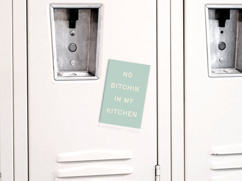 The Onderkast Studio No Bitchin’ In My Kitchen Refrigerator Magnet in mint green decorates a white locker, boldly displaying its message in uppercase—perfect kitchen decor and part of a locker row.